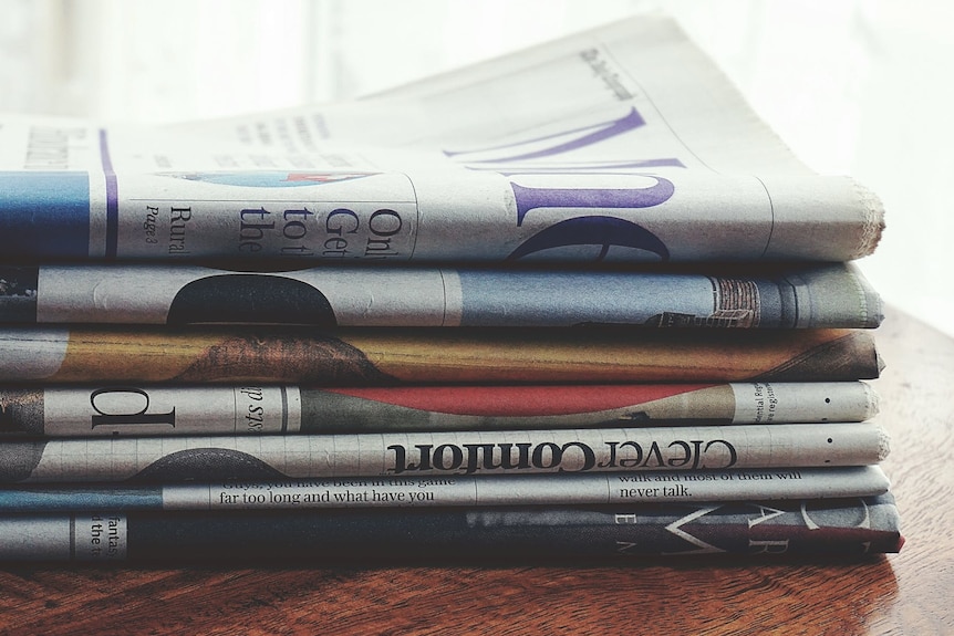 A stack of seven English language newspapers are seen on a wooden table, folded.