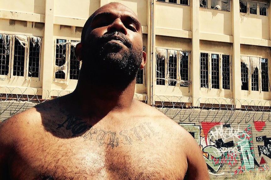 A young shirtless man stands in front of a wall with graffiti and broken windows.