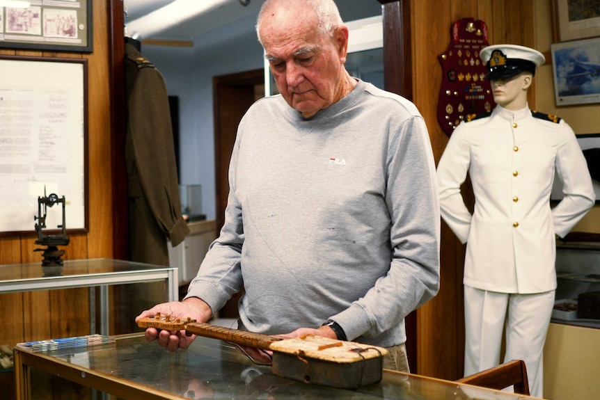 A man examines a small, wooden, stringed instrument on a glass bench with a mannequin in naval uniform behind him.