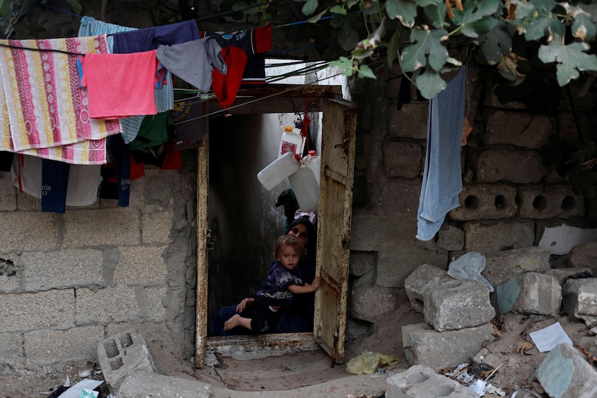A woman holding a child sits in the doorway of a home made out of cinder blocks. More blocks and laundry surround the entryway.