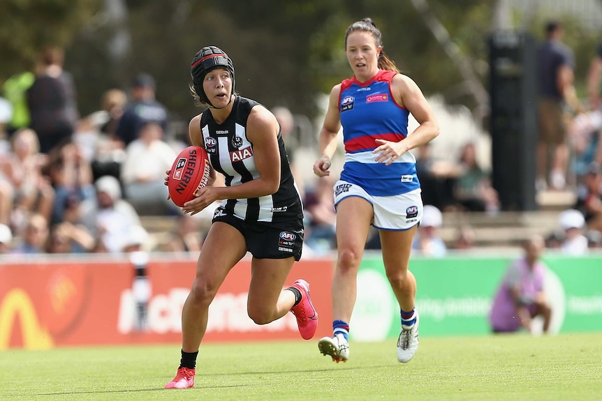 An AFLW player wearing headgear looks up as she runs with the ball as an opponent chases her.