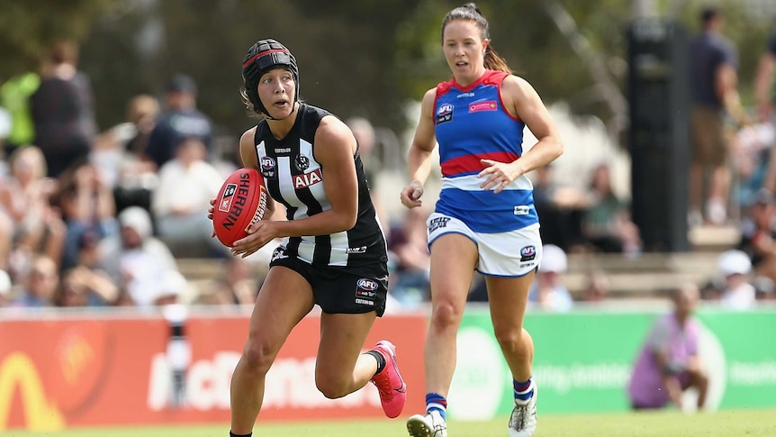 An AFLW player wearing headgear looks up as she runs with the ball as an opponent chases her.