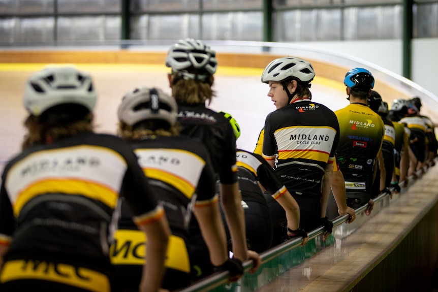 Teenagers lined up holding on to railing on their bicycles in a velodrome.