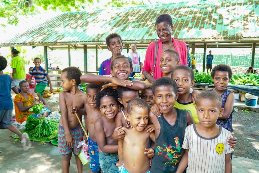 A smiling woman in a red top looks down on a group of children posing for a photo