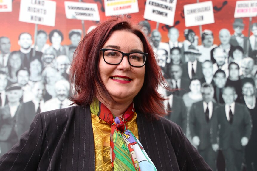 A woman with red hair, a colourful scarf and a black blazer stands in front of a mural.