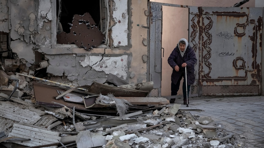 Elderly woman stands near her destroyed home.