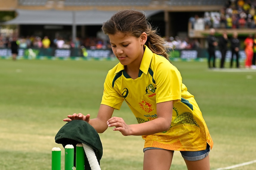 A young girl places a baggy green on the stumps.