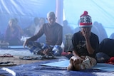 Two villagers sit in a temporary smoky shelter in Bali one man in the foreground has his hand to his face