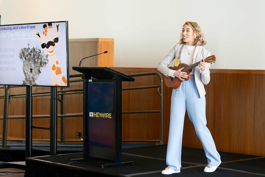 Young woman on stage holding a guitar with a screen behind her with a cute animation of a giraffe and a yak.