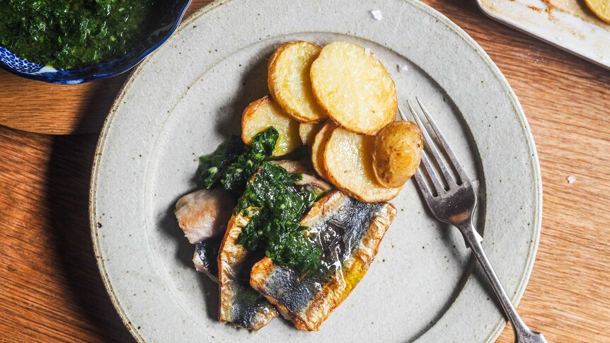 A plate of dinner with three butterflied sardines topped with parsley sauce and slices or roast potato, a seafood dinner.