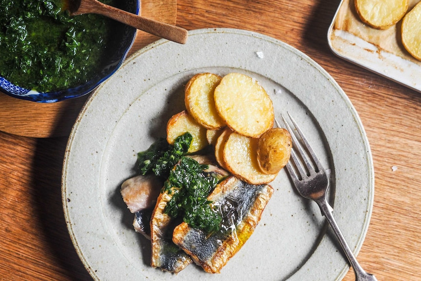 A plate of dinner with three butterflied sardines topped with parsley sauce and slices or roast potato, a seafood dinner.