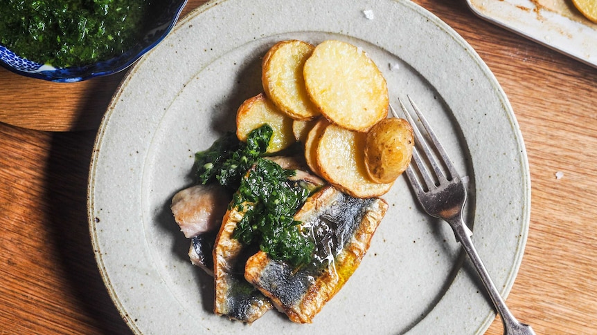 A plate of dinner with three butterflied sardines topped with parsley sauce and slices or roast potato, a seafood dinner.