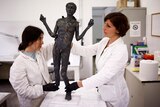 Two young white women in white lab coats hold up on a lab table a roughly metre-long, dark-coloured statue of a skinny boy.