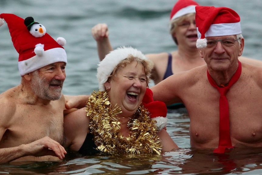 Two men wearing Santa hats stand in the water and are either side of a laughing woman dressed in tinsel.