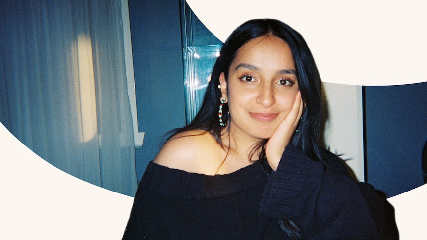 A woman is seen looking to the camera and slightly smiling, her hand resting on her cheek. Her dark hair is down.