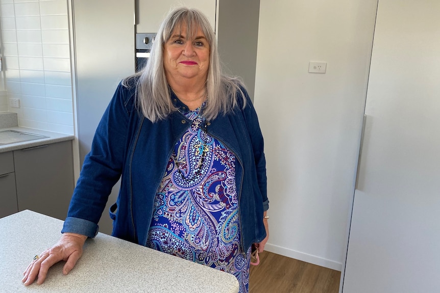 Woman with long silver hair and a purple top stands in an empty kitchen.