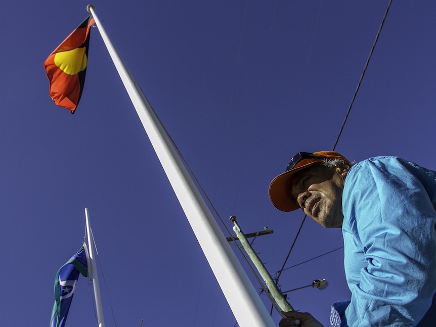 An Indigenous man raises the Aboriginal and Torres Strait Island flags.