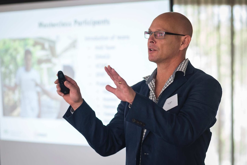 A bald man with glasses talks in front of a Powerpoint presentation.