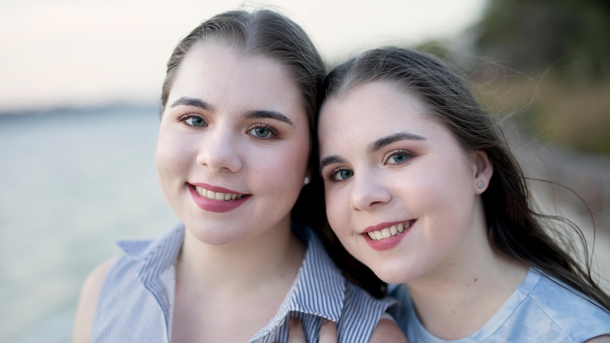 Two identical twins women,  wearing blue tops at a beach have their heads together and smile at the camera.