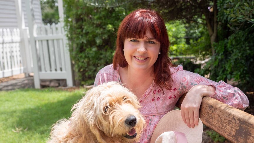 Leah Stirton with red hair and a pink dress pats her dog and smiles to the camera.