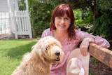 Leah Stirton with red hair and a pink dress pats her dog and smiles to the camera.