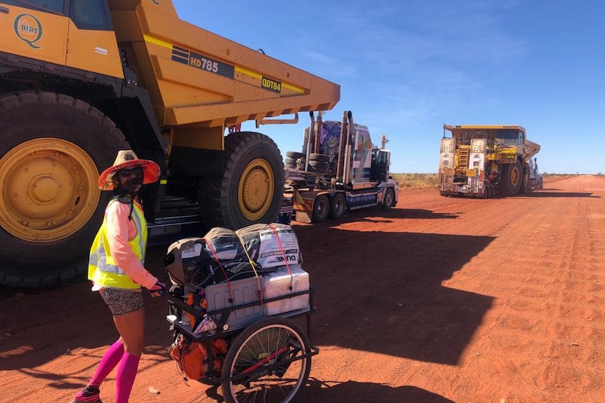 A woman pushing a hand cart is dwarfed by huge mining dump trucks on a sandy road.