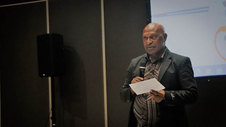 A man stands in front of a screen, speaking into microphone while holding a page of notes