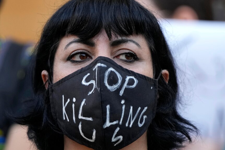 A woman at a protest wears a black face mask with the words 'Stop Killing Us' drawn onto it.