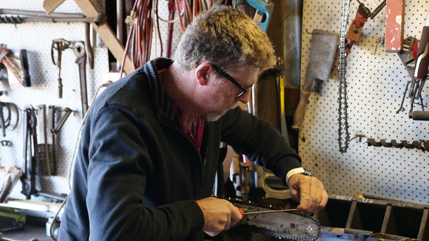 A man wearing glasses in a tool shed sharpens his chainsaw