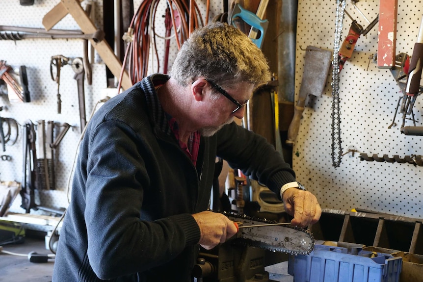A man wearing glasses in a tool shed sharpens his chainsaw