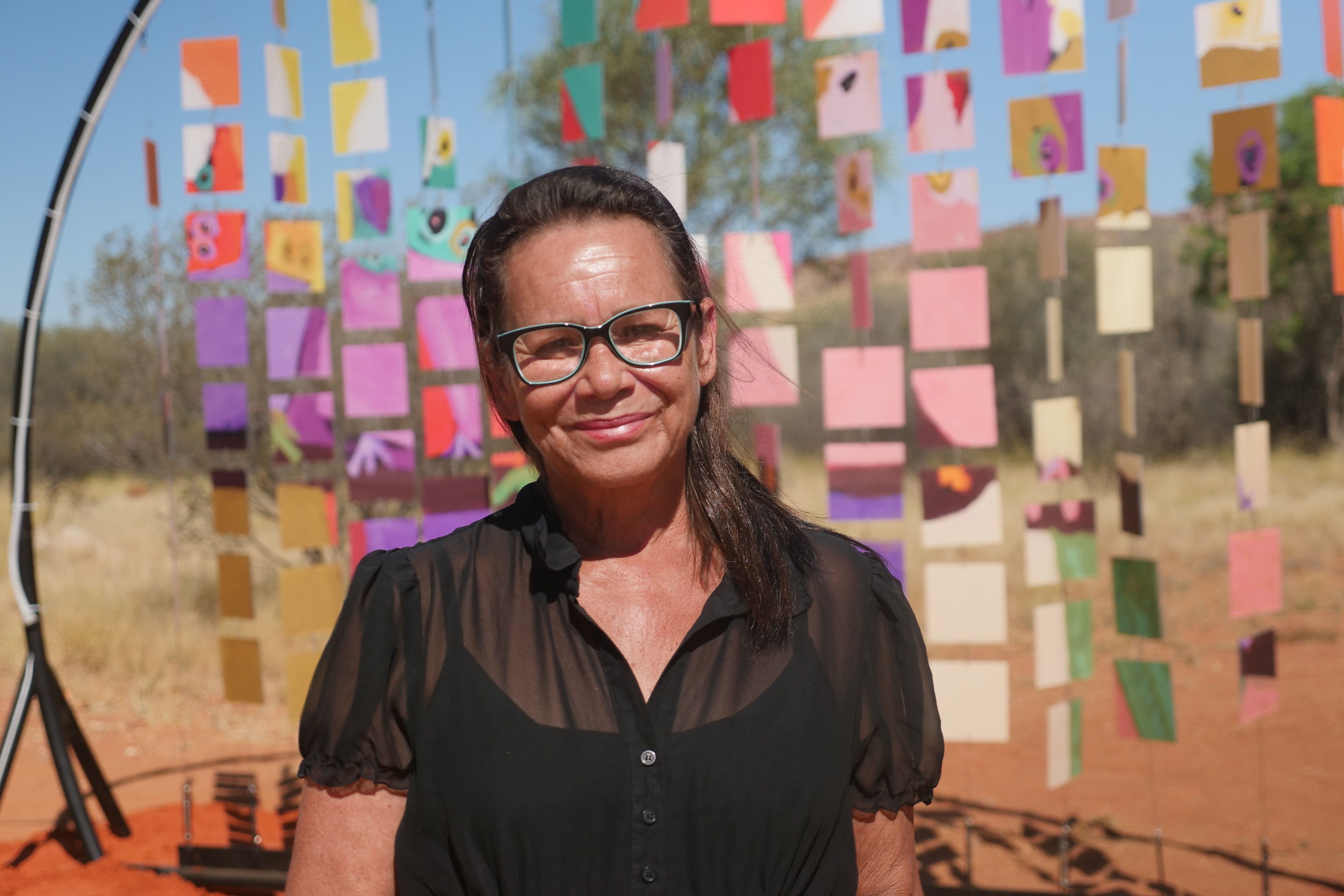 An Indigenous woman wearing glasses stands in front of a colourful outback art installation, she is smiling at the camera.