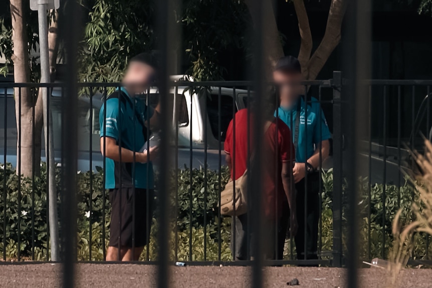 Two men in black caps and blue shirts talk to a person on the street. The photo is shot from a distance through bars.
