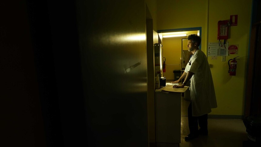 A doctor wearing a white coat is silhouetted in a room as he looks at a bright screen showing X-rays.