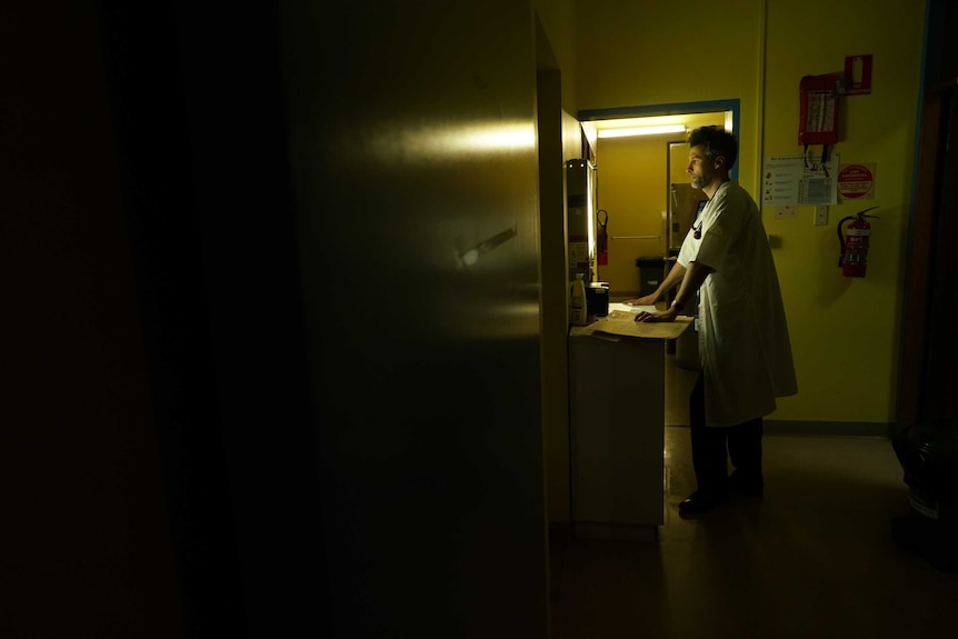 A doctor wearing a white coat is silhouetted in a room as he looks at a bright screen showing X-rays.