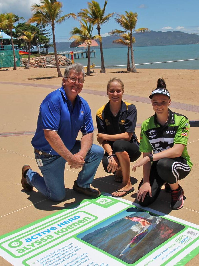 One man and two young female athletes pose alongside a large sticker on Townsville's foreshore walk