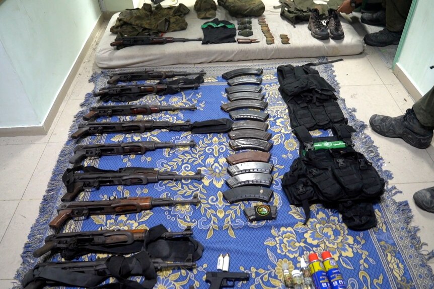 Israeli soldiers stand near an array of rifles, magazines, body armour and other weapons laid out on a blue and gold rug.