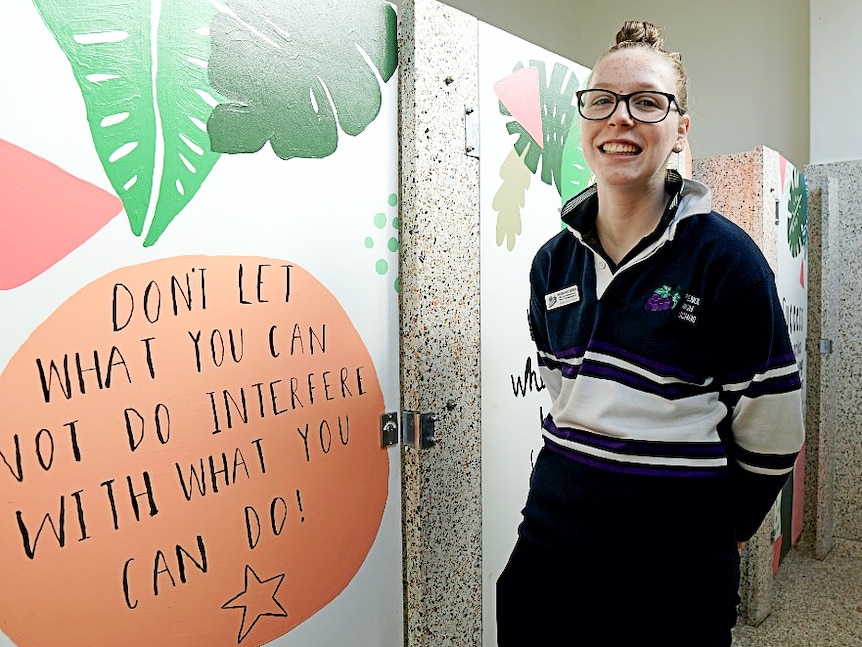 Brooke Hutchinson stands in front of door with the words 'Don't let what you can not do interfere with what you can do' painted.
