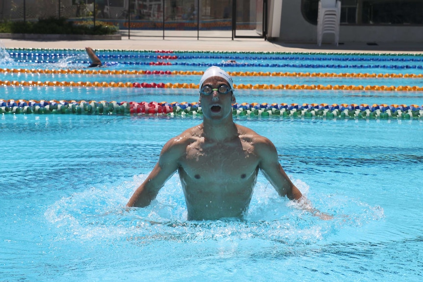 Ethan Calleja emerges from the water in a pool with his waist and arms above the surface, wearing a swimming cap and goggles.