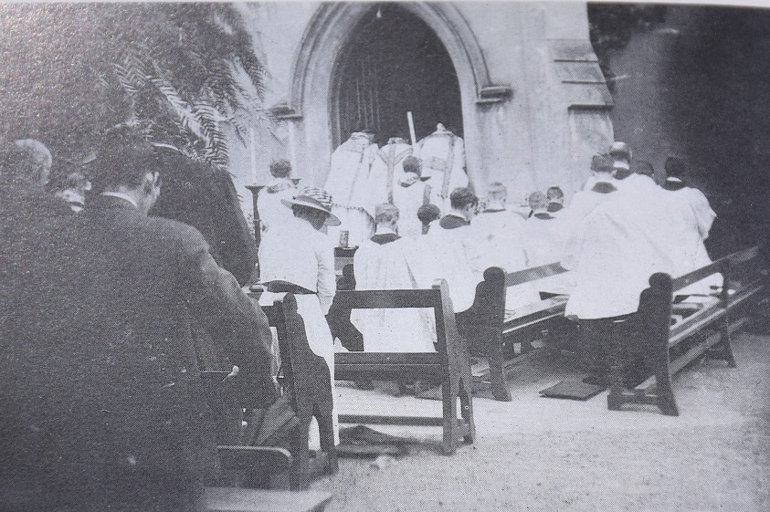 A black and white photograph shows a congregation meeting for a service outdoors.