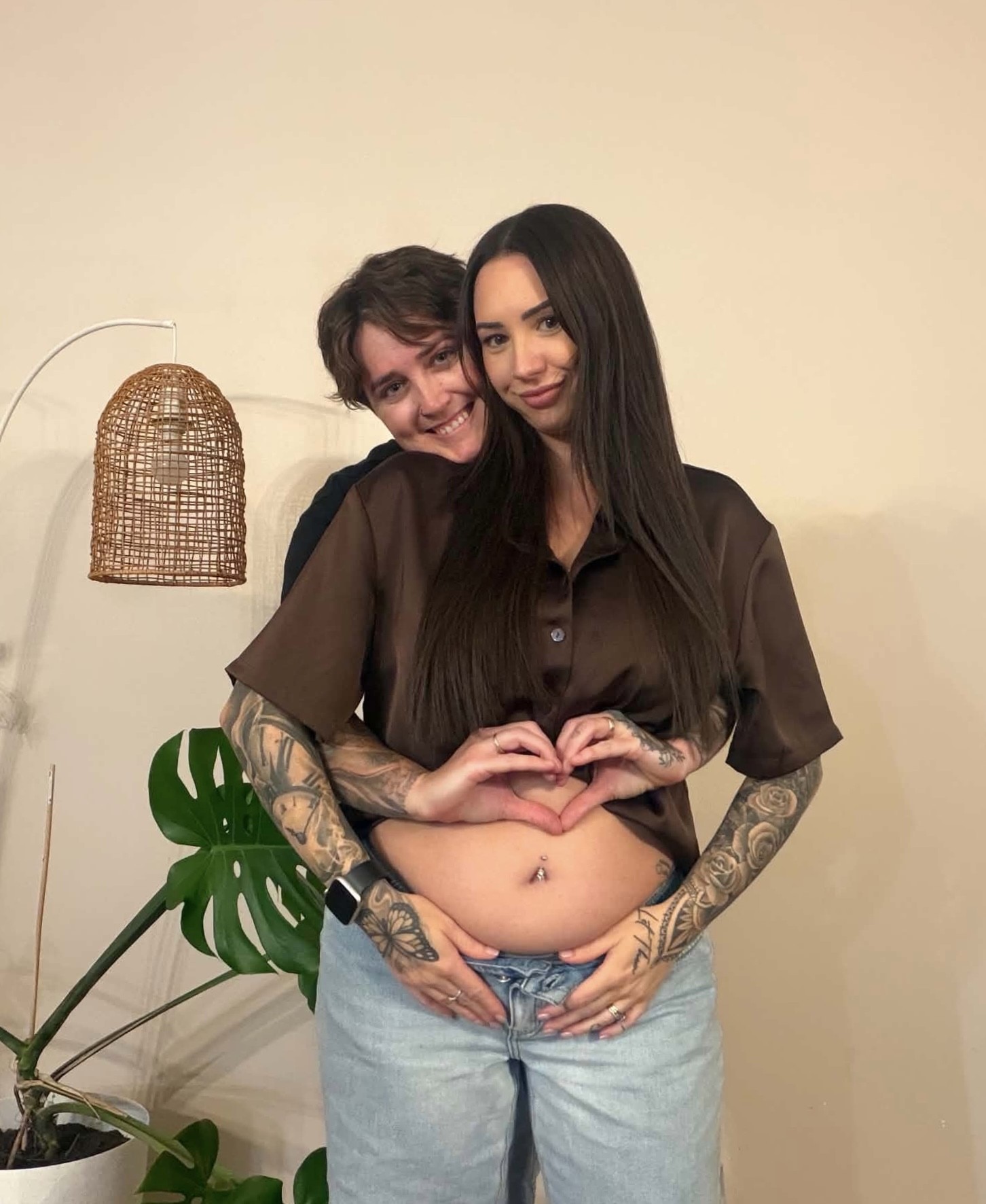 A woman standing behind her female partner making a heart shape with her hands above her partner's belly.