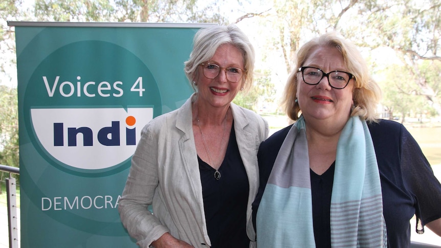 Two women with glasses and fair hair stand next to a green banner that says 'Voices 4 Indi'.