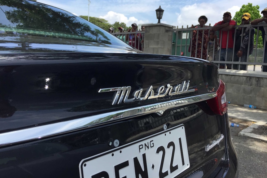 a black car bearing PNG number plates in front of a fence with people behind it