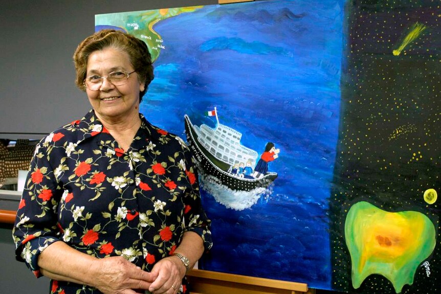 A smiling Gina Sinozich stands in front of an artwork depicting a woman in a boat.