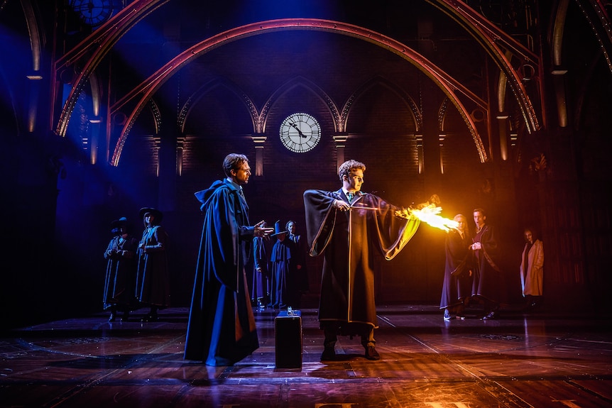 Two actors in long black cloaks perform on a gothic-style stage. One is holding a wand that shoots fire.