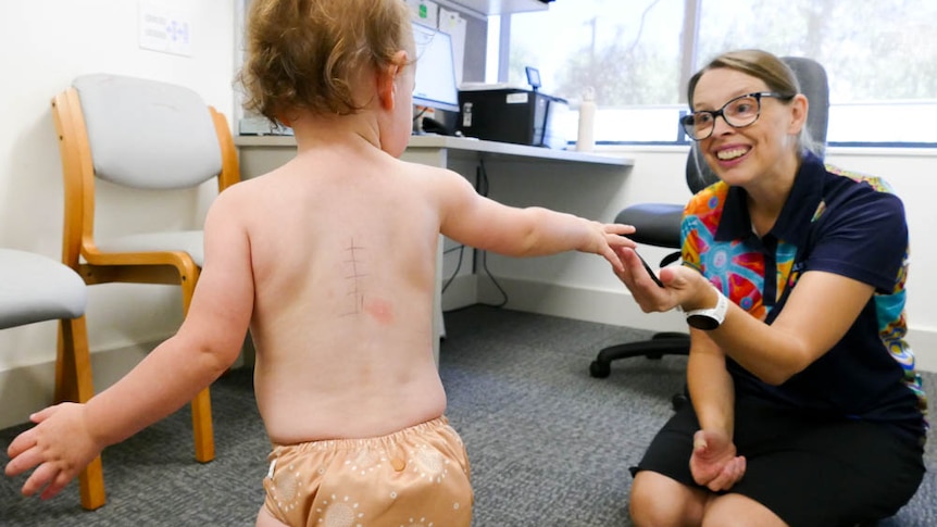 A baby with cross marks on its back approaches a smiling woman sitting on her knees