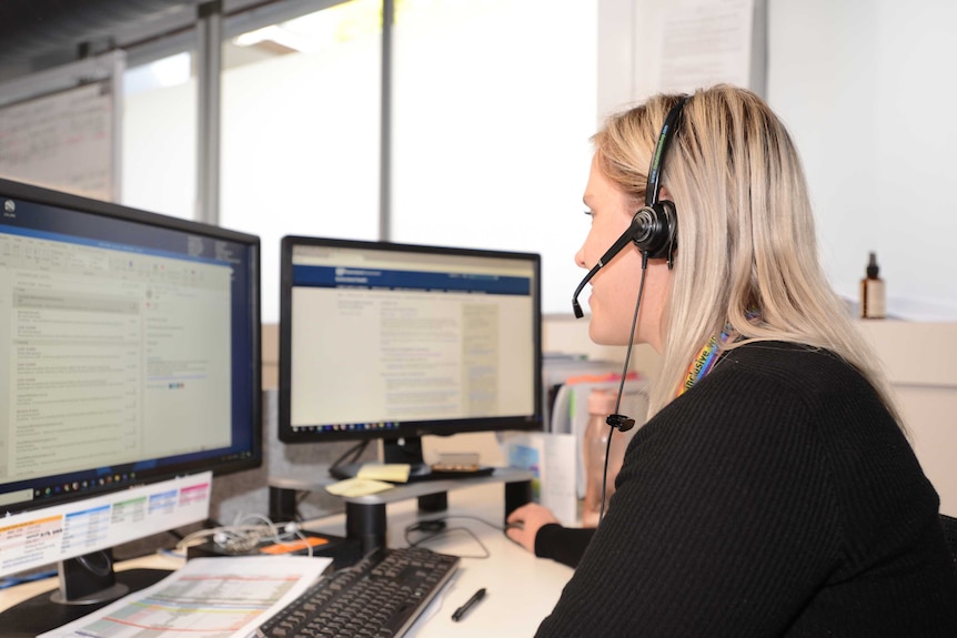 A blonde woman sits facing computer screens and talking into a headset.