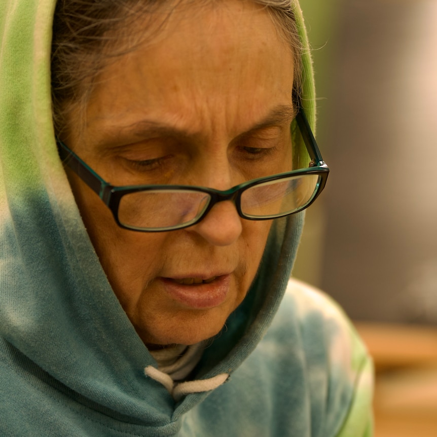 A middle aged white woman with black glasses wearing a rainbow hoodie. She is sitting at a table making art