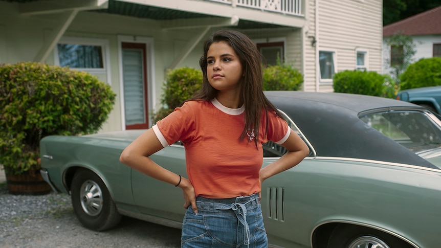 A young woman wearing denim shorts and orange t-shirt stands with hands on hips in front of vintage car and suburban house.