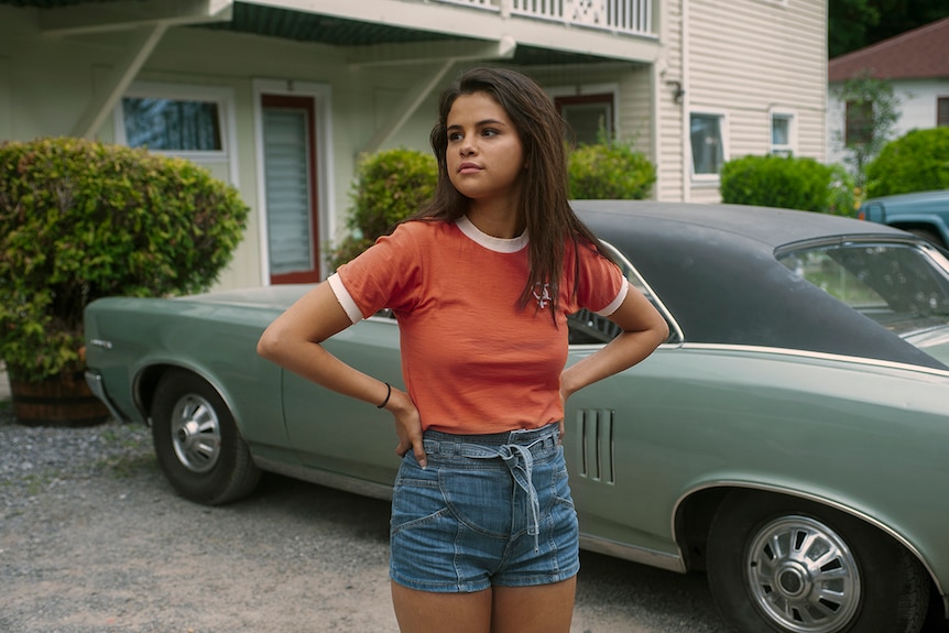 A young woman wearing denim shorts and orange t-shirt stands with hands on hips in front of vintage car and suburban house.