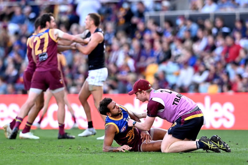 Callum Ah Chee is attended to by a medic while lying on the ground. Behind him, two Lions players wrestle Patrick Cripps